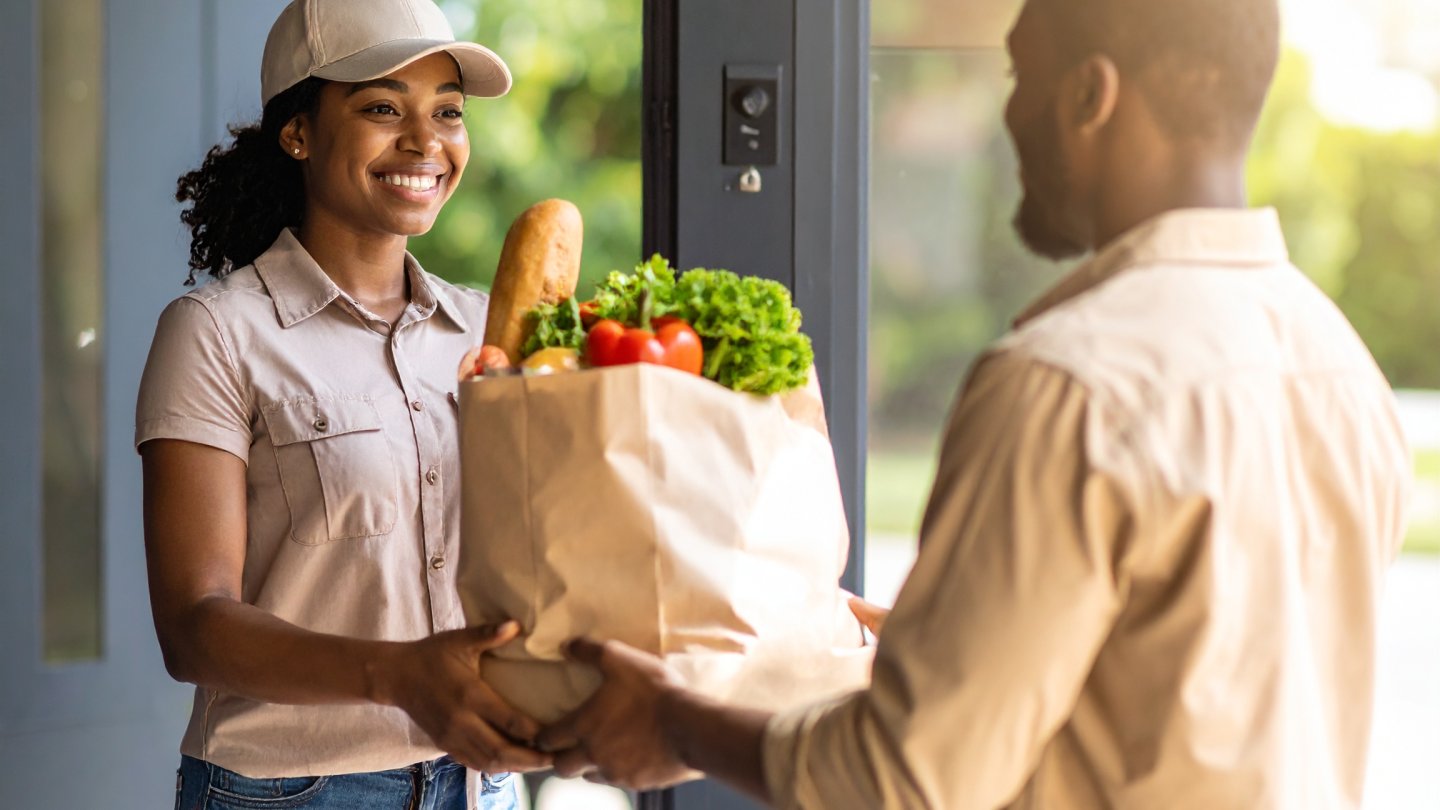 A woman handing a grocery delivery to a man at his doorstep, showcasing the convenience and efficiency of home delivery services.